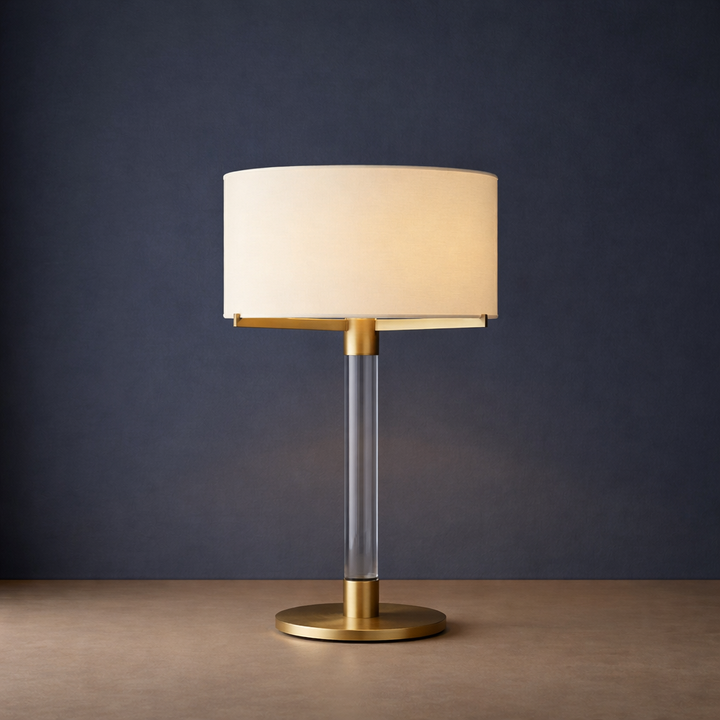 Moro aged brass metal table lamp featuring a cylindrical linen shade, clear glass column stem, and brushed brass base in a refined minimalist setting